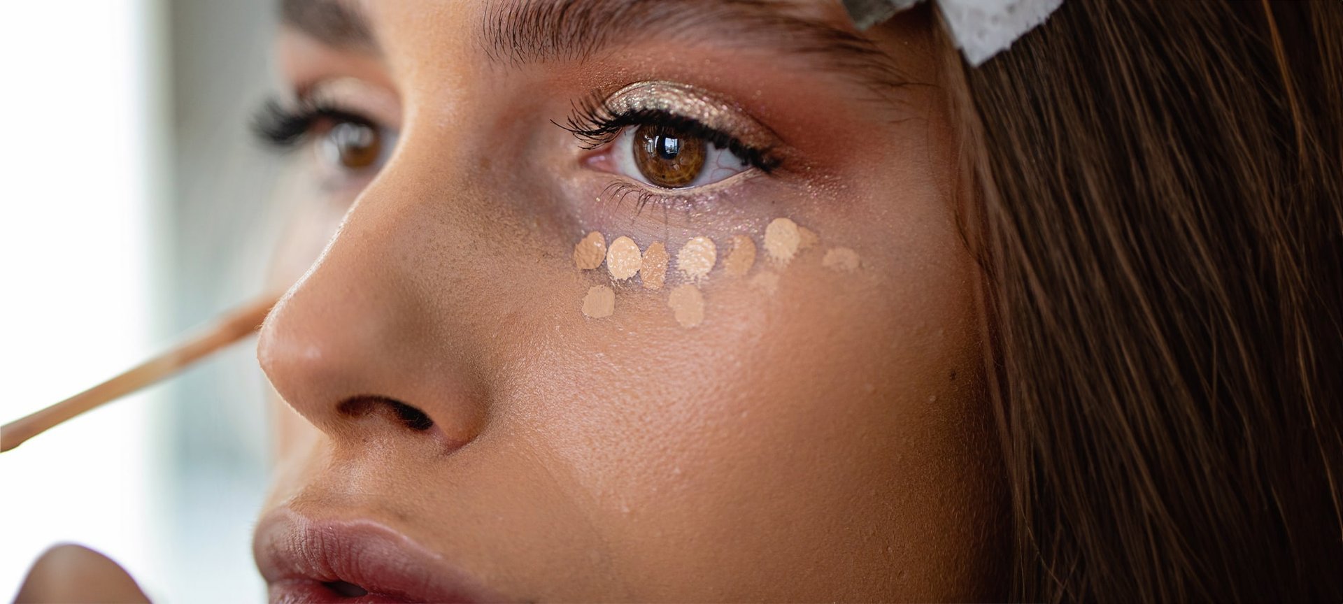 Junge, geschminkte Frau trägt unter ihren Augen Concealer in Tupfenform auf.