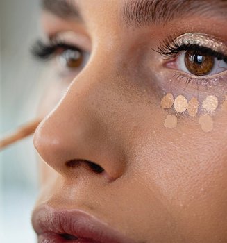 Junge, geschminkte Frau trägt unter ihren Augen Concealer in Tupfenform auf.