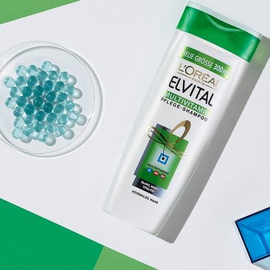 Petrischale mit blauen Kügelchen liegt neben Elvital Multivitamin Shampoo und einem blauen, gelben und roten Würfel.