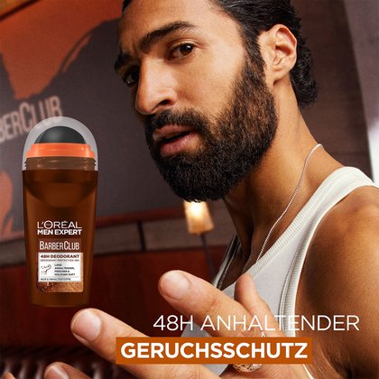 Eine Hand haelt das Barber Club Bartoel vor roter Steinmauer