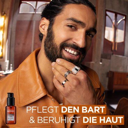 Ein lächelnder Mann mit dunklem Bart und einer braunen Lederjacke berührt sein Kinn. Unten links ist das Barber Club Bartöl abgebildet. Text im Bild: „PFLEGT DEN BART & BERUHIGT DIE HAUT“
