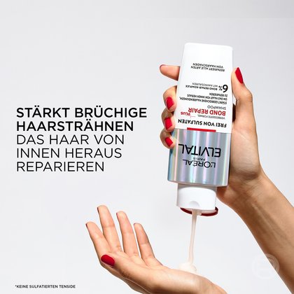 Eine Hand mit rot lackierten Fingernägeln hält die Shampooflasche über Kopf und gibt eine Portion weißes, cremiges Shampoo auf die Handfläche einer zweiten Hand. Links steht der Text: „STÄRKT BRÜCHIGE HAARSTRÄHNEN. DAS HAAR VON INNEN HERAUS REPARIEREN.*“ mit dem Hinweis „*Keine sulfatierten Tenside“.