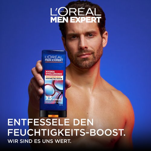 Ein Mann mit nasser Haut hält die dunkelblaue Flasche des „Hydra Hyaluronic Duschgels“ direkt in die Kamera. Oben links ist das weiße Logo von „L'Oréal Men Expert“ zu sehen; darunter steht der Slogan: „ENTFESSELE DEN FEUCHTIGKEITS-BOOST. WIR SIND ES UNS WERT.“
