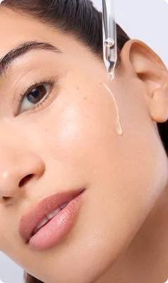 Gesicht einer jungen Frau mit hellem Hautton; Serumflüssigkeit tröpfelt aus einer Pipette auf die Wange der Frau