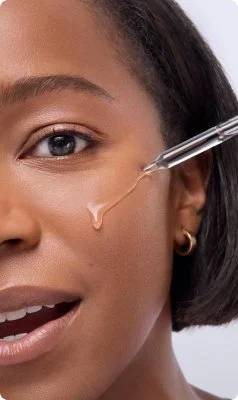 Gesicht einer jungen Frau mit dunklem Hautton; Serumflüssigkeit tröpfelt aus einer Pipette auf die Wange der Frau