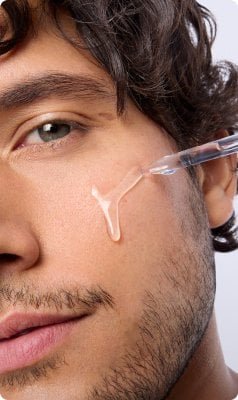 Gesicht eines Mannes mit mittlerem Hautton und Bart; Serumflüssigkeit tröpfelt aus einer Pipette auf die Wange des Mannes