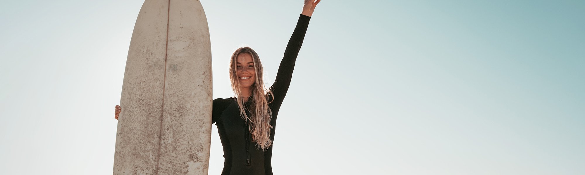 Junge Frau mit blonden Haaren am Strand mit einem Surfbrett in der Hand