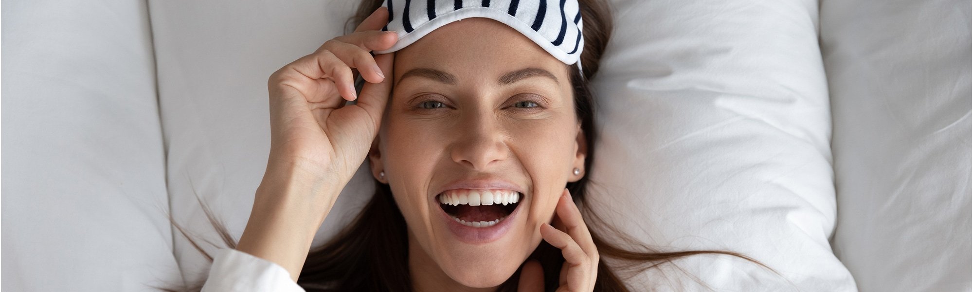 Junge Frau liegt lächelnd im Bett mit Schlafmaske auf der Stirn