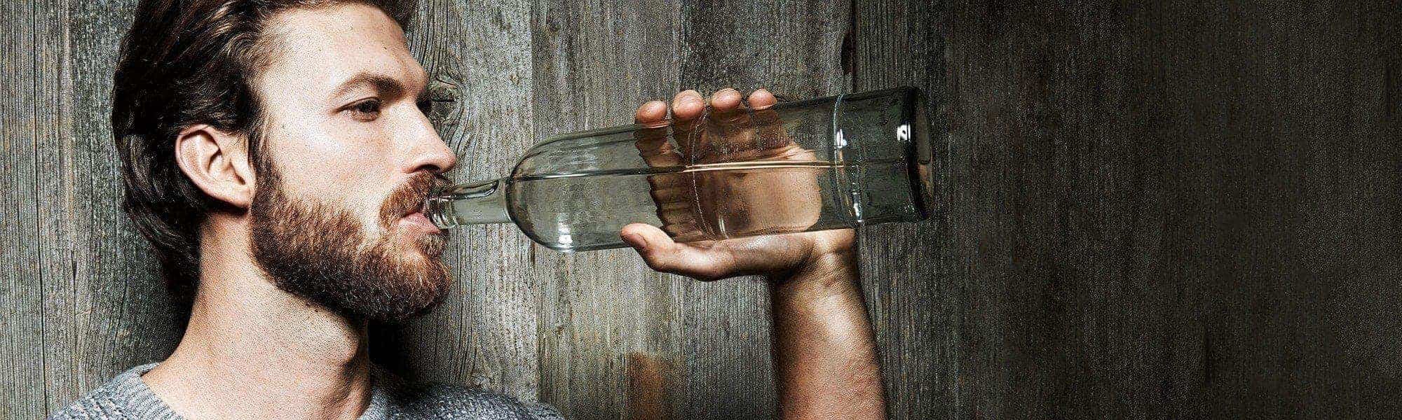 Bärtiger Mann trinkt aus Wasserfalsche vor einer Holzwand